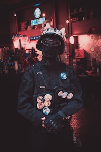 A person dressed in a dark tactical outfit stands in a dimly lit interior space. They are wearing a helmet with cat ears, goggles, and a black face covering. The outfit is adorned with various buttons featuring anime characters. The background appears to be a decorated room with soft lighting, shelves, and a neon sign.