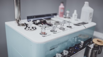A collection of various dispensing and control devices on a white platform with a metallic rod structure. There are bottles of different shapes and sizes, likely containing liquid solutions, and several small glass containers. The surface is equipped with multiple nozzles and handles.