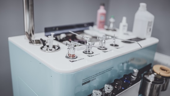 A collection of various dispensing and control devices on a white platform with a metallic rod structure. There are bottles of different shapes and sizes, likely containing liquid solutions, and several small glass containers. The surface is equipped with multiple nozzles and handles.