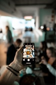 A person holds a smartphone, capturing a photo of a table set with various dishes and a group of people in a dining setting. The focus is on the smartphone screen, which shows the food and company more clearly than the blurred background.