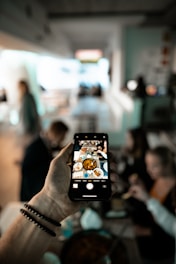 A person holds a smartphone, capturing a photo of a table set with various dishes and a group of people in a dining setting. The focus is on the smartphone screen, which shows the food and company more clearly than the blurred background.