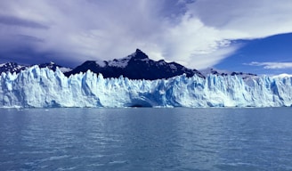 A large, expansive glacier with jagged ice formations spans across the horizon. The glacier is backed by steep, dark mountain peaks under a partly cloudy sky. The water in the foreground reflects the icy blue and white hues of the glacier.