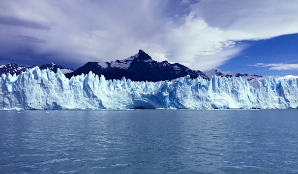 A large, expansive glacier with jagged ice formations spans across the horizon. The glacier is backed by steep, dark mountain peaks under a partly cloudy sky. The water in the foreground reflects the icy blue and white hues of the glacier.