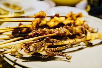 Several skewers loaded with grilled meat, possibly including squid and other types of seafood, are placed on a plate. The skewers appear to be charred and seasoned, indicating a freshly grilled and appetizing presentation. The background is slightly blurred, focusing on the skewers in the foreground.