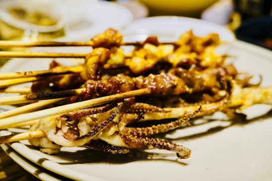 Several skewers loaded with grilled meat, possibly including squid and other types of seafood, are placed on a plate. The skewers appear to be charred and seasoned, indicating a freshly grilled and appetizing presentation. The background is slightly blurred, focusing on the skewers in the foreground.