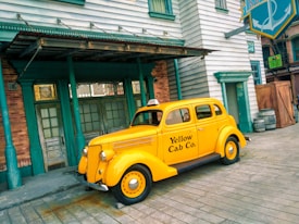 A vintage yellow taxi with the text 'Yellow Cab Co.' on the side is parked on a wooden street in front of a building with green trim and large windows. The scene has a nostalgic feel, with elements reminiscent of a bygone era.