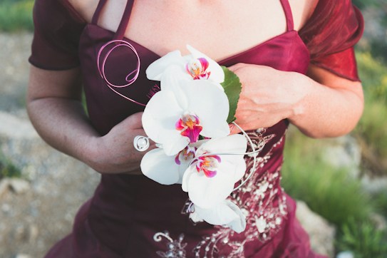 A person wearing a deep burgundy dress holds a bouquet of white orchids. The dress features intricate embroidery and the orchids have vibrant pink and yellow centers, adding a touch of elegance. The setting appears to be outdoors with a blurred natural background.