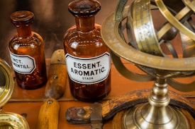 Several vintage brown bottles with labels, including one labeled 'ESSENT. AROMATIC,' are seen alongside a brass armillary sphere with engraved markings. The background includes wooden handles and a surface with a warm hue.