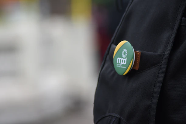 A close-up view of a backpack with a pinned badge on it. The badge has a green and yellow design, featuring a white text logo 'mpd' accompanied by a graphic of interconnected circles. The badge appears to be attached to a brown leather strap on the backpack. The background is blurred, with indistinct light colors suggesting an outdoor environment.