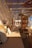 A cozy outdoor seating area lined with cushions and pillows in various patterns and colors. The area is shaded by a wooden pergola with a woven cover, casting intricate shadows. Natural materials and soft lighting create a warm and inviting atmosphere.