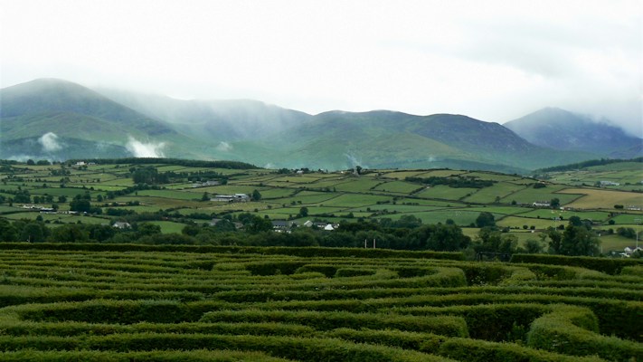 A lush, green landscape featuring neatly trimmed hedges in the foreground, with rolling hills and farmland in the middle ground. Misty mountains are visible in the background, partially covered by low clouds.