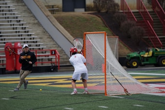 Two individuals are on a sports field, equipped with lacrosse sticks. One appears to be a goalie in front of the net, while the other is positioned to the side, possibly in preparation to take a shot. The field is marked with lines and features a net behind the goalie. In the background, there are empty bleachers and training equipment.