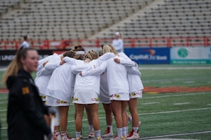 A group of athletes dressed in white uniforms is huddled together on a sports field, displaying team unity and preparing for a game. The stadium seating in the background is mostly empty, creating a focused atmosphere on the team.