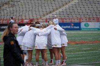 A group of athletes dressed in white uniforms is huddled together on a sports field, displaying team unity and preparing for a game. The stadium seating in the background is mostly empty, creating a focused atmosphere on the team.