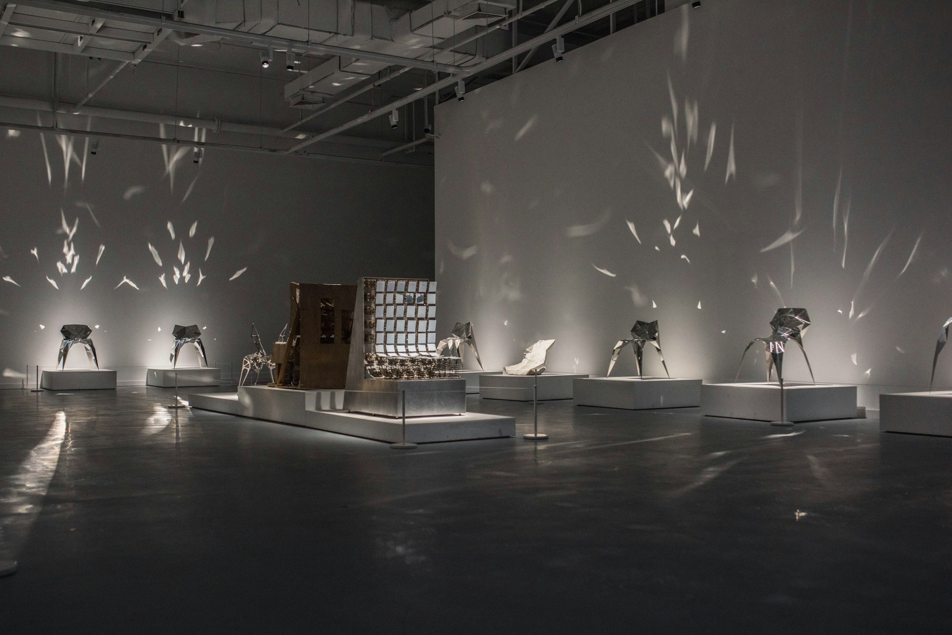 An art gallery installation featuring modern, abstract sculptures exhibited on individual platforms. The room is dimly lit with focused lighting that casts intricate shadow patterns on the walls. The sculptures have metallic and mirrored surfaces, reflecting light in various directions.