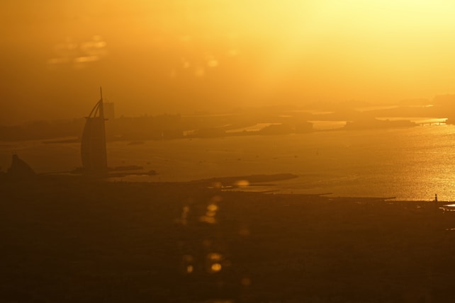 A golden sunset over a cityscape by the sea with a prominent sail-shaped building silhouetted against the vibrant sky. The sun casts a warm, golden hue across the water, creating an almost ethereal atmosphere.