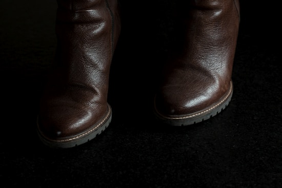 A pair of brown leather boots with a rugged texture and a visible zipper on the side. The boots have sturdy soles with treads suitable for outdoor use. The lighting casts a shadow, highlighting the polished surface of the leather.