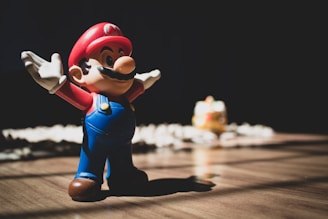 A small, toy figurine of a character wearing a red hat and blue overalls is standing on a wooden surface. The figurine has large white gloves, a mustache, and is posed with arms wide open. Soft light casts a shadow behind the toy. In the background, there is a blurred object that resembles another toy.