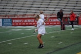 A girl in sports attire, wearing goggles, is practicing lacrosse on an outdoor field. She is holding a lacrosse stick, and the background features empty stadium bleachers and a banner promoting women's lacrosse championships. Several other individuals are in the background, possibly coaches or team members.