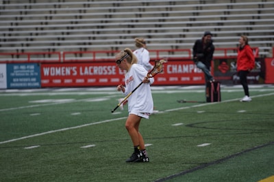 A girl in sports attire, wearing goggles, is practicing lacrosse on an outdoor field. She is holding a lacrosse stick, and the background features empty stadium bleachers and a banner promoting women's lacrosse championships. Several other individuals are in the background, possibly coaches or team members.