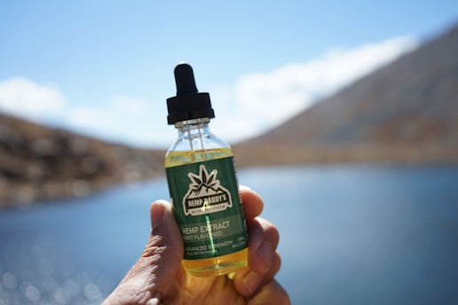 A hand is holding a small bottle labeled 'Hemp Daddy’s Therapeutics' containing hemp extract with a dropper cap. The background shows a blurred outdoor setting with clear blue sky and bodies of water, suggesting a lake or pond area.