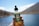 A hand is holding a small bottle labeled 'Hemp Daddy’s Therapeutics' containing hemp extract with a dropper cap. The background shows a blurred outdoor setting with clear blue sky and bodies of water, suggesting a lake or pond area.