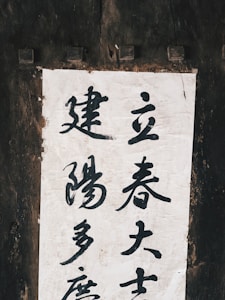 A piece of paper with Chinese calligraphy is mounted on a rustic, dark wooden surface. The calligraphy is done in black ink, featuring multiple characters vertically arranged on a light, textured background.