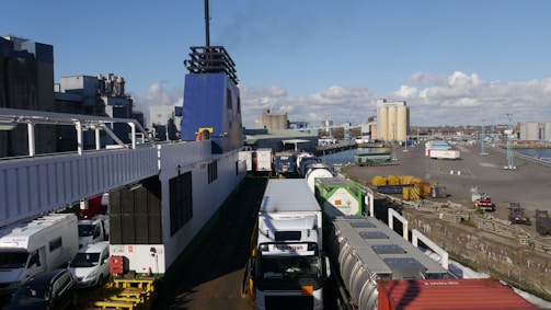 A busy industrial port scene featuring a large ferry docked at the harbor. Numerous trucks and trailers are lined up, ready for loading or unloading. The background shows industrial buildings and storage tanks, with a clear blue sky and scattered clouds.