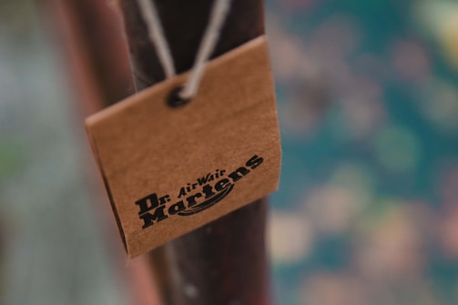A close-up of a brown tag with the Dr. Martens logo, attached to a pole with a string. The background is blurred with soft tones.