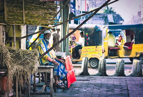 A man wearing a colorful striped shirt and a white cap is sitting on a wooden stool beside a stack of sugarcane and straw. In the background, there are bright yellow auto rickshaws on a busy street. The scene has an urban market feel with various street elements visible.