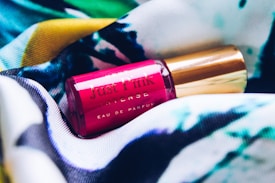 A small pink perfume bottle labeled 'Just Pink Intense' rests on a multicolored fabric. The fabric features a mix of blue, green, yellow, and white hues with abstract patterns.