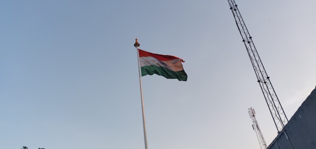 A tall flagpole with the Indian national flag waving prominently against a clear sky, accompanied by a tall metal tower and small silhouettes of structures on the ground.