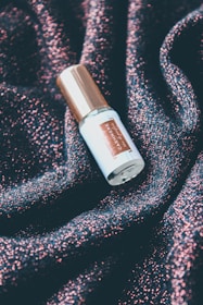 A bottle with a metallic gold cap labeled with the word 'cashmere'. It is resting on a textured surface that sparkles with shades of pink and dark blue, resembling a glittery fabric.
