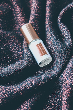 A bottle with a metallic gold cap labeled with the word 'cashmere'. It is resting on a textured surface that sparkles with shades of pink and dark blue, resembling a glittery fabric.