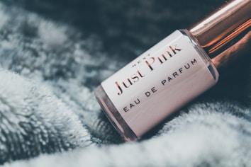 A close-up view of a perfume bottle labeled 'Just Pink Eau de Parfum' resting on a soft, fluffy surface. The bottle has a rose gold cap and a sleek, cylindrical shape, exuding an elegant and feminine aesthetic.