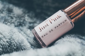 A close-up view of a perfume bottle labeled 'Just Pink Eau de Parfum' resting on a soft, fluffy surface. The bottle has a rose gold cap and a sleek, cylindrical shape, exuding an elegant and feminine aesthetic.