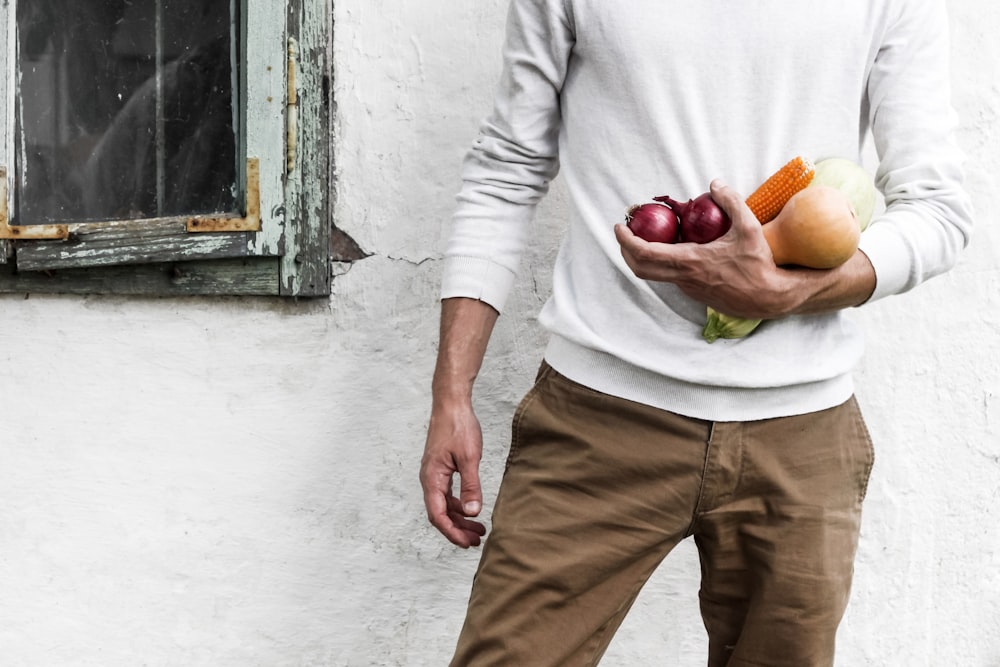 man standing beside white wall while holding fruits