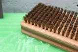 A close-up view of a rectangular wooden brush with densely packed, medium-bristle fibers. The bristles appear slightly worn and are arranged in neat rows on the wooden base. The background consists of a textured green surface, possibly plastic or painted, with a slightly blurred gray wall in the distance.