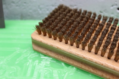 A close-up view of a rectangular wooden brush with densely packed, medium-bristle fibers. The bristles appear slightly worn and are arranged in neat rows on the wooden base. The background consists of a textured green surface, possibly plastic or painted, with a slightly blurred gray wall in the distance.
