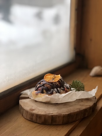 A pastry topped with chocolate drizzle and a dried orange slice sits on a piece of parchment paper. It rests on a rustic wooden cutting board by the window, with a sprig of evergreen as garnish. The window is slightly fogged, hinting at a cold or wintry atmosphere outside.