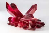 A cluster of vibrant red crystals with translucent facets is arranged on a light background. The crystals have a glossy and polished appearance, reflecting light and displaying different shades of red along their surfaces.