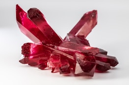 A cluster of vibrant red crystals with translucent facets is arranged on a light background. The crystals have a glossy and polished appearance, reflecting light and displaying different shades of red along their surfaces.