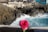 A vibrant pink rose in the foreground contrasts with the rugged coastline and crashing waves in the background. The rocky shore is bordered by a construction area with multiple unfinished pillars and small buildings. The ocean water is a bright turquoise hue.