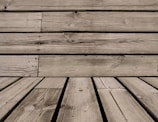 Wooden planks with visible grain patterns and some knots, arranged horizontally and vertically. The wood has a weathered appearance, with natural imperfections and variations in color.