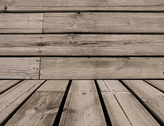 Wooden planks with visible grain patterns and some knots, arranged horizontally and vertically. The wood has a weathered appearance, with natural imperfections and variations in color.