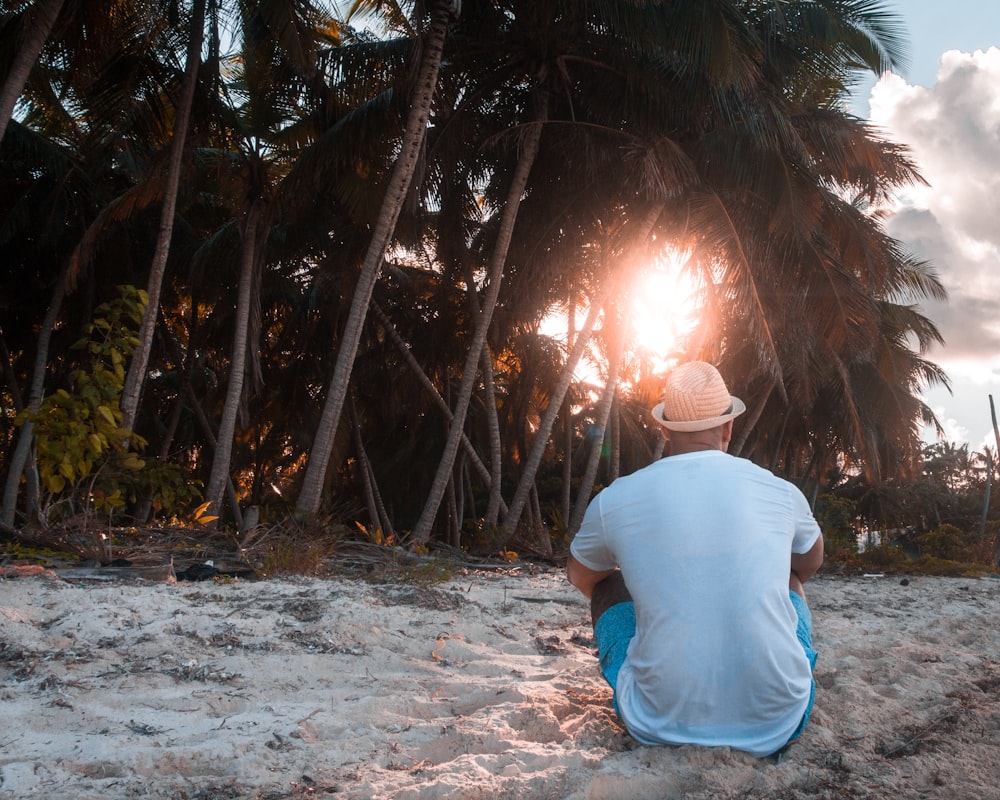 man in white shirt and brown hat sitting on sand facing coconut trees
