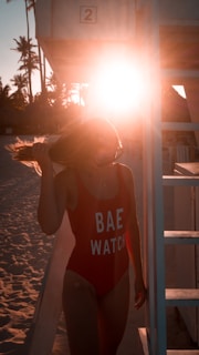 A person in a red swimsuit with the text 'BAE WATCH' stands next to a lifeguard tower on a sandy beach. The sun is setting, casting a warm glow over the scene, and palm trees are visible in the background.