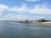 A coastal scene features several quaint houses and a lighthouse along a sandy shoreline. The calm, blue ocean gently laps against the shore under a wide, partly cloudy sky. Small boats can be seen floating on the water, adding to the serene atmosphere of the setting.