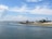 A coastal scene features several quaint houses and a lighthouse along a sandy shoreline. The calm, blue ocean gently laps against the shore under a wide, partly cloudy sky. Small boats can be seen floating on the water, adding to the serene atmosphere of the setting.