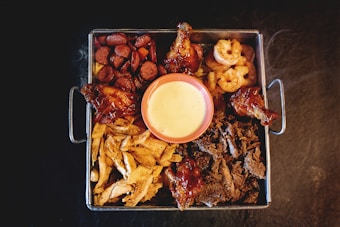 A rectangular metal tray filled with various grilled and roasted food items surrounds a central bowl of pale yellow dipping sauce. The tray contains sausage slices, chicken wings, shrimp, shredded beef, and slices of chicken breast. The arrangement is visually appetizing with a mix of textures and colors.